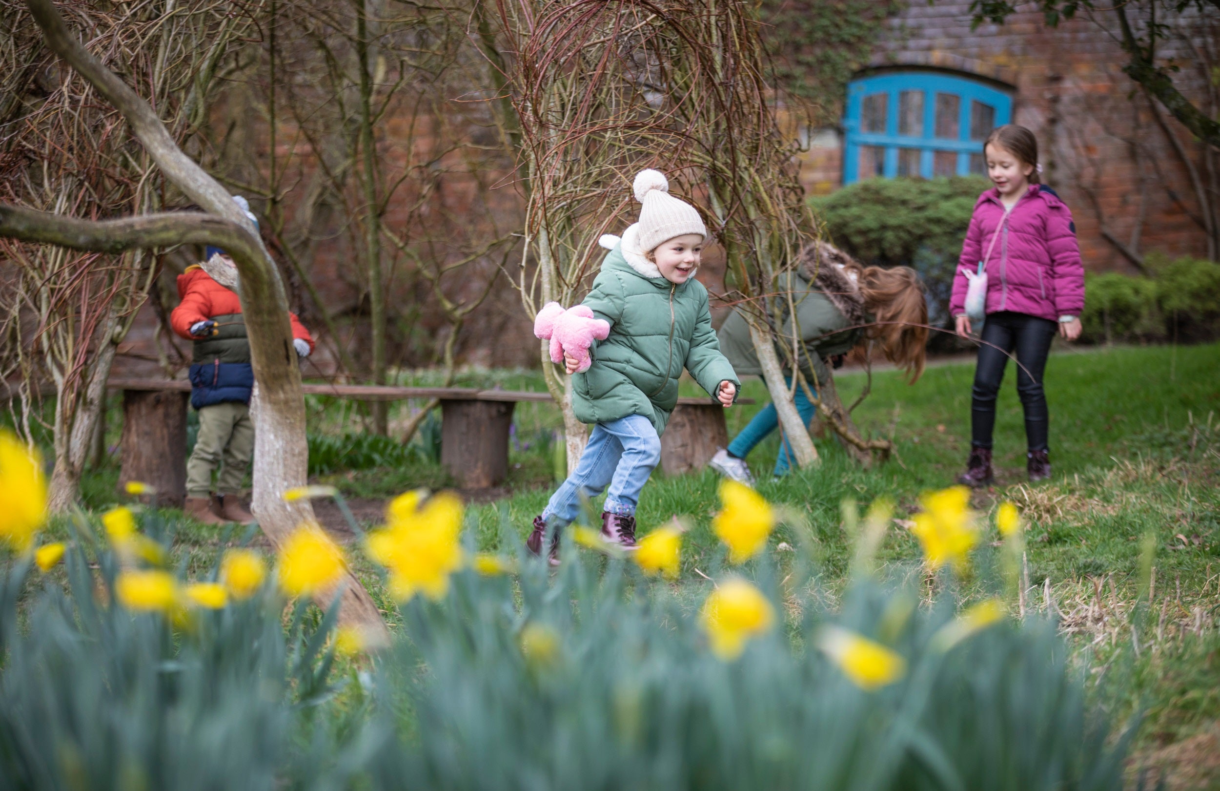 Child running, holding a teddy bear, with a background of trees and daffodils in the front.