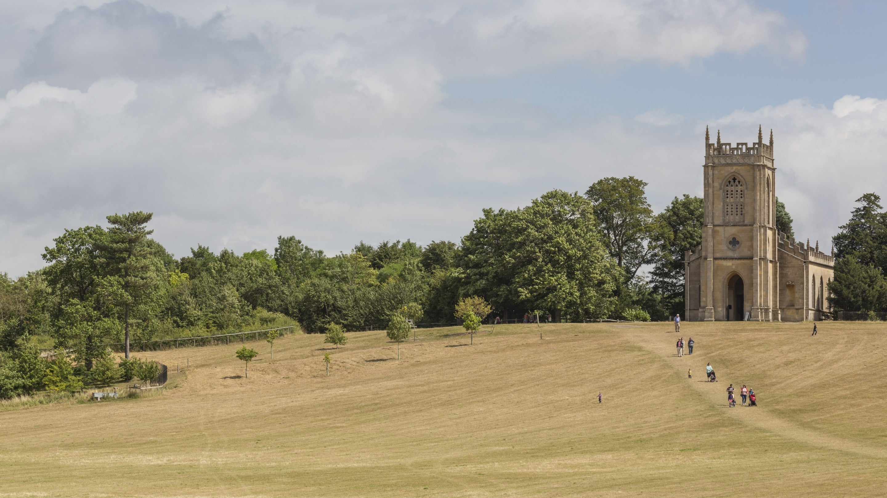 The Gothic church of St Mary Magdalene at Croome, Worcestershire, created by Capability Brown sits on a hill. Visitors walk up the hill across grass faded yellow by the sun. Trees line the horizon behind the church.