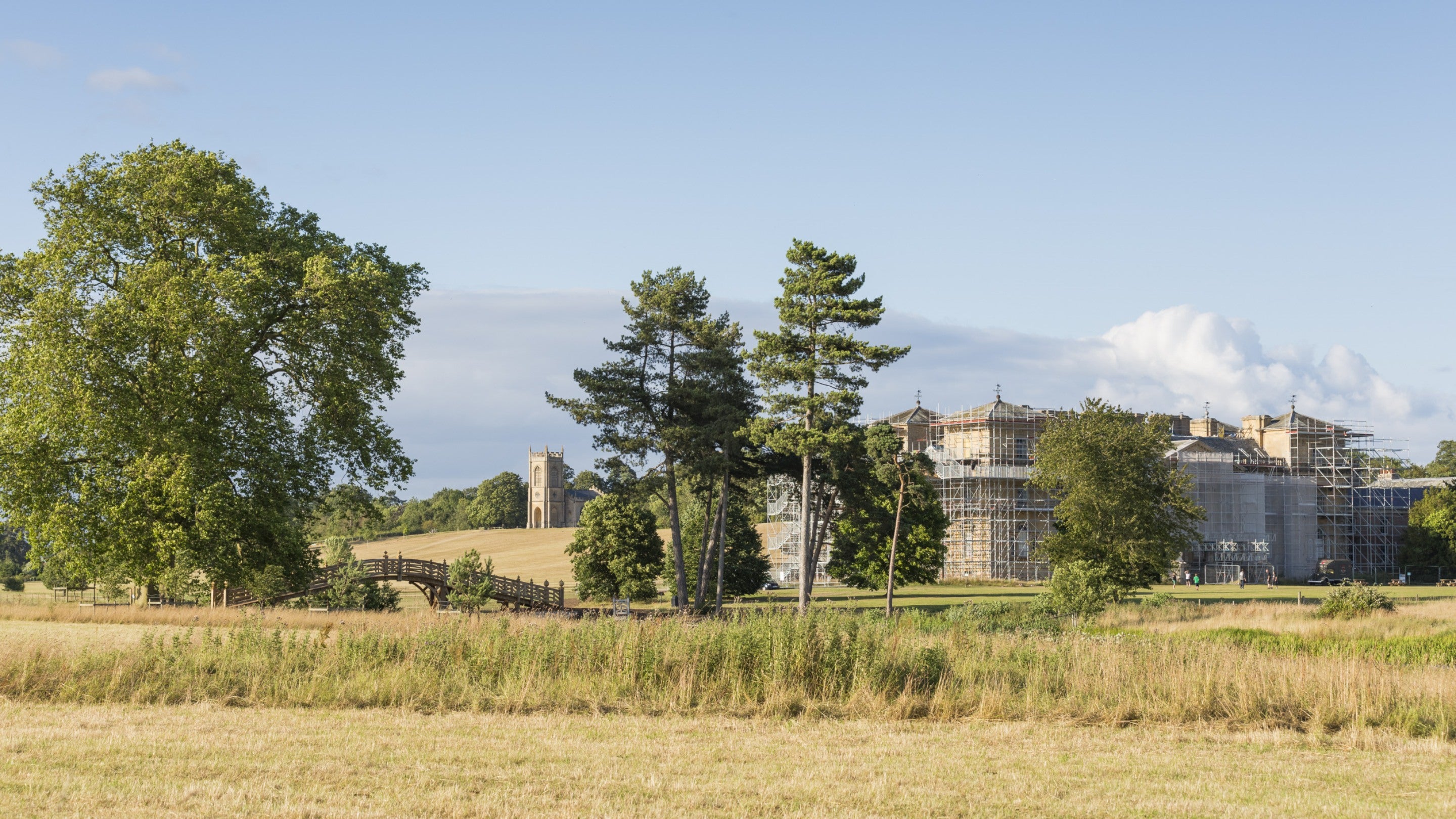A view across the landscape towards the house at Croome, which is covered in scaffolding, with trees in the foreground and the house and church in the distance