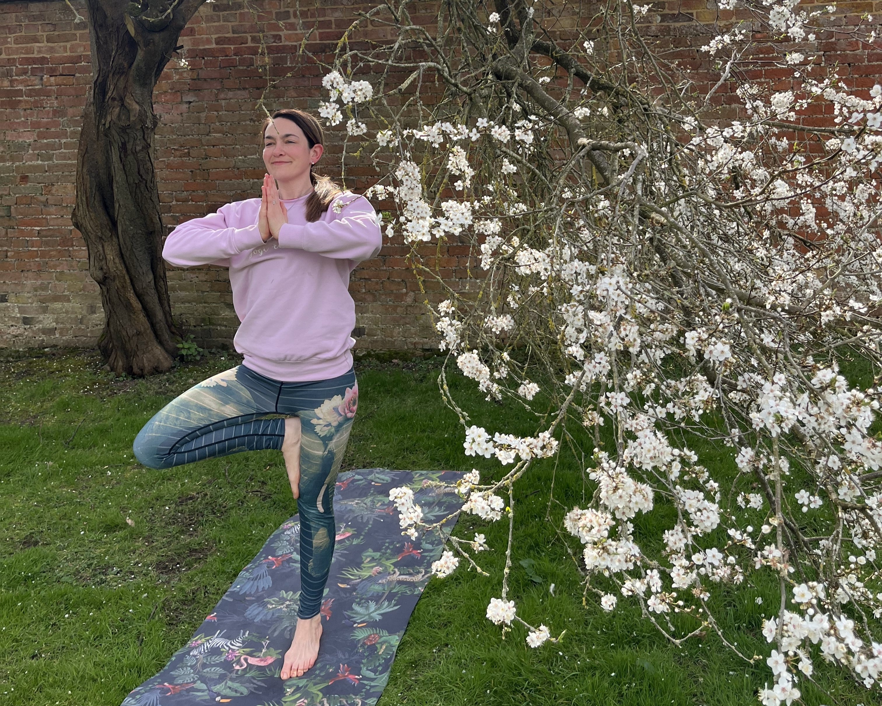 A yoga teacher doing a tree yoga pose on a yoga mat underneath a blossom tree in the orchard