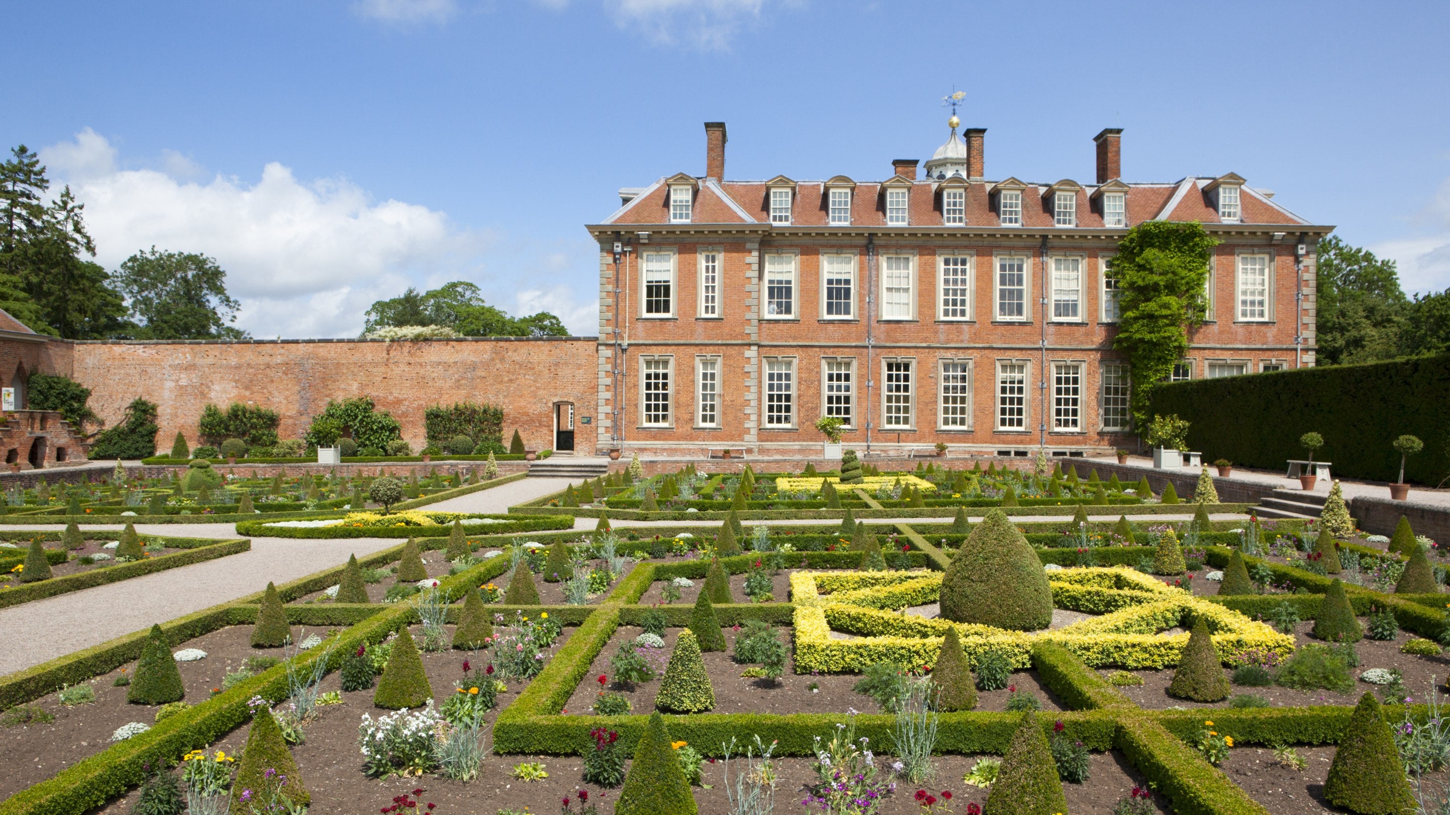The restored parterre at Hanbury Hall, Worcestershire with neatly clipped box hedging in a geometric pattern interspersed with summer flowers. Behind the parterre the red brick exterior of the house can be seen.
