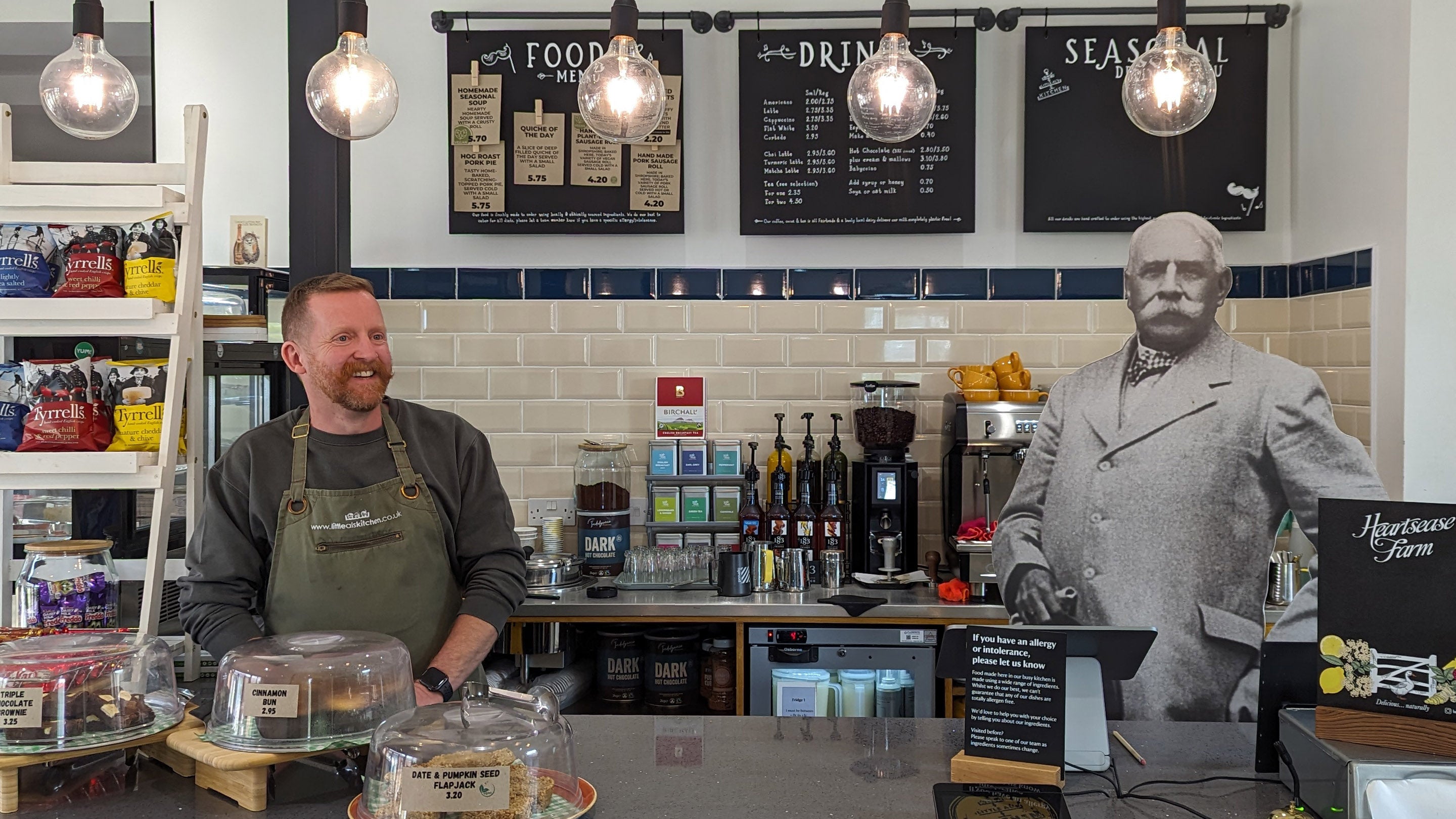 The counter at Little Al's cafe at The Firs showing Little Al smiling with a life-size cut-out of Edward Elgar surrounded by coffee, tea and cakes