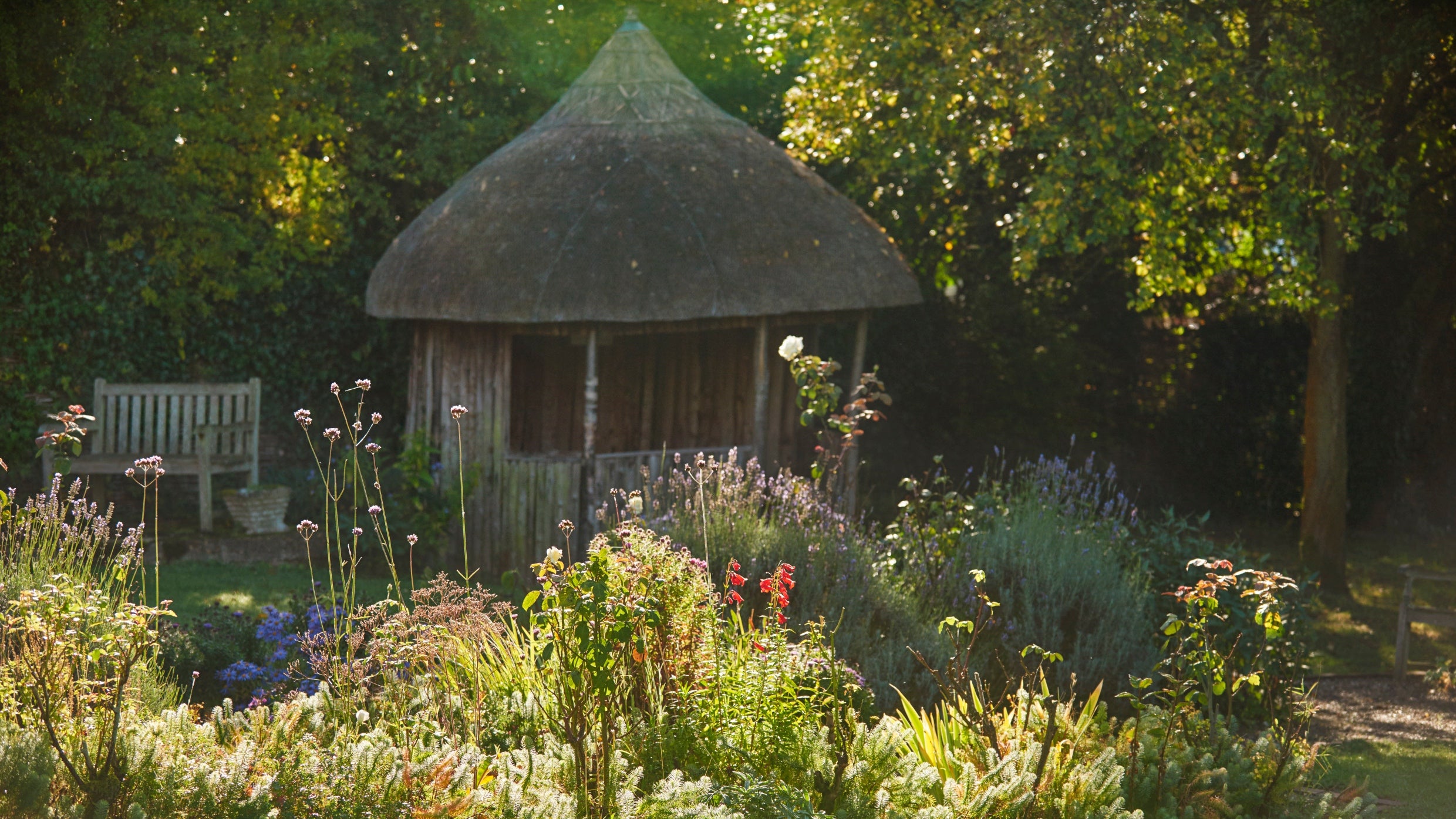 The Summerhouse, a thatched wooden hut in the garden behind a bed of wildflowers