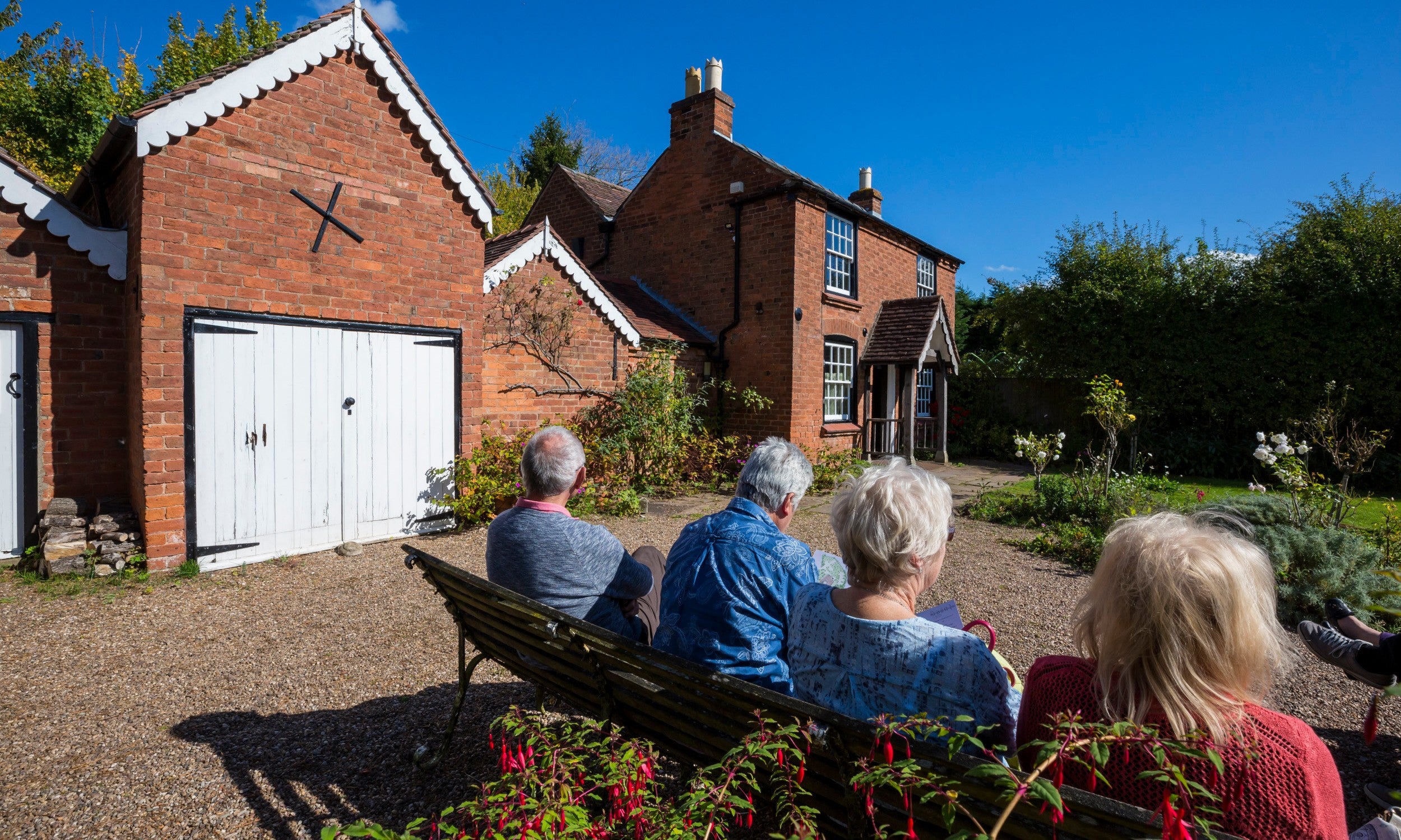 Group of four elderly visitors sitting on a bench in the garden at The Firs, facing the house