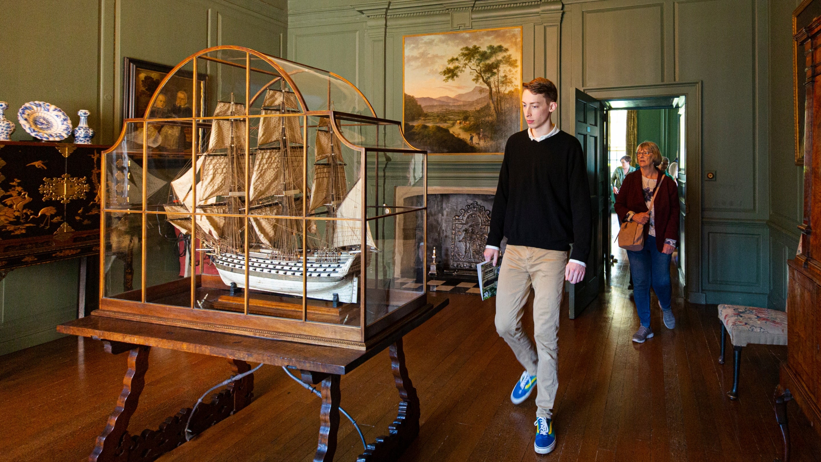 A large model ship with intricate sails and guns detail inside a glass cabinet on display at Treasurers House. Two visitors are walking into the dark wood floor and green walled room to look at it.