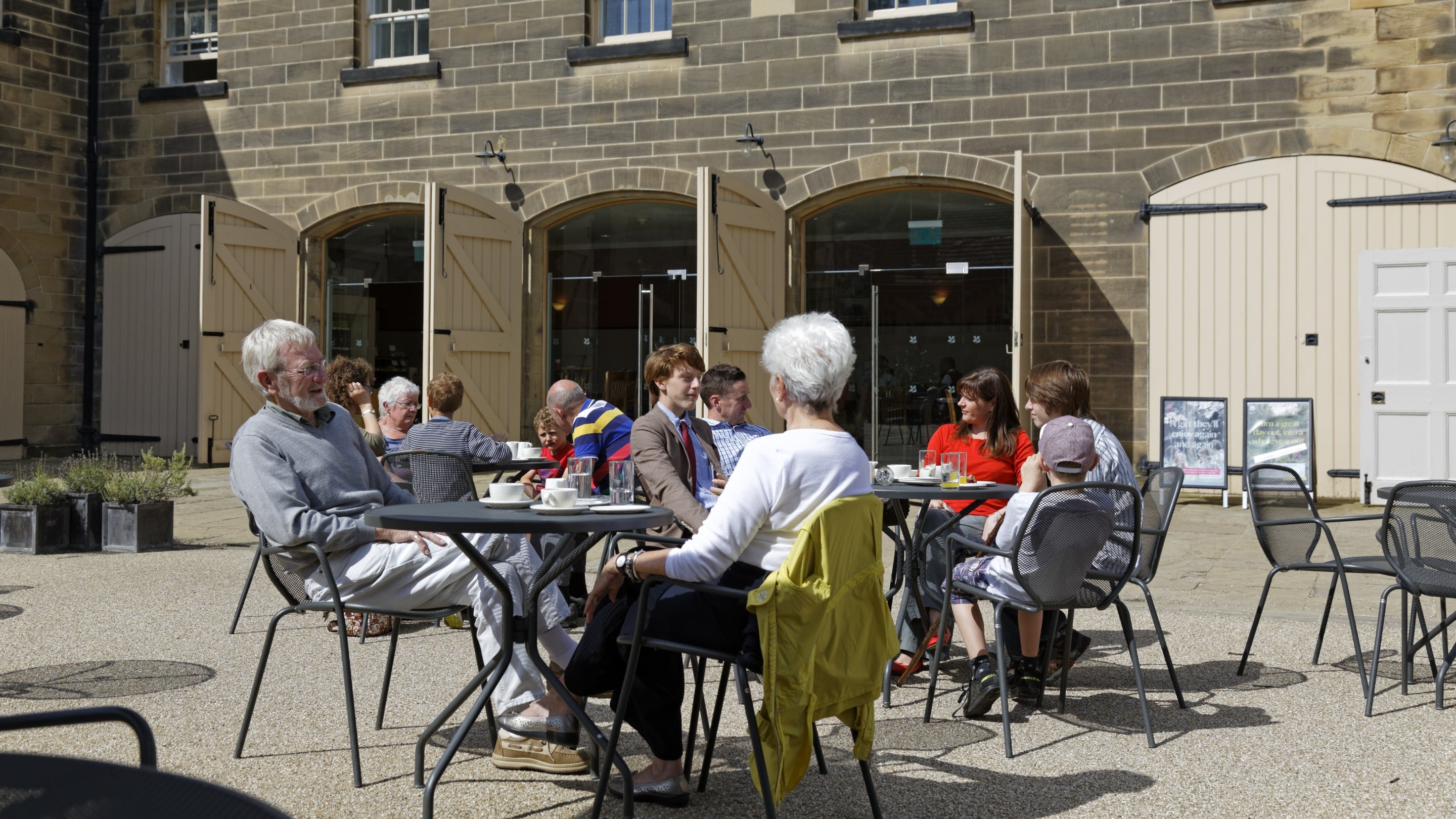 People are sat outside at tables and chairs, you can see drinks and some food on the table. Behind them is the Stables Cafe with its doors open