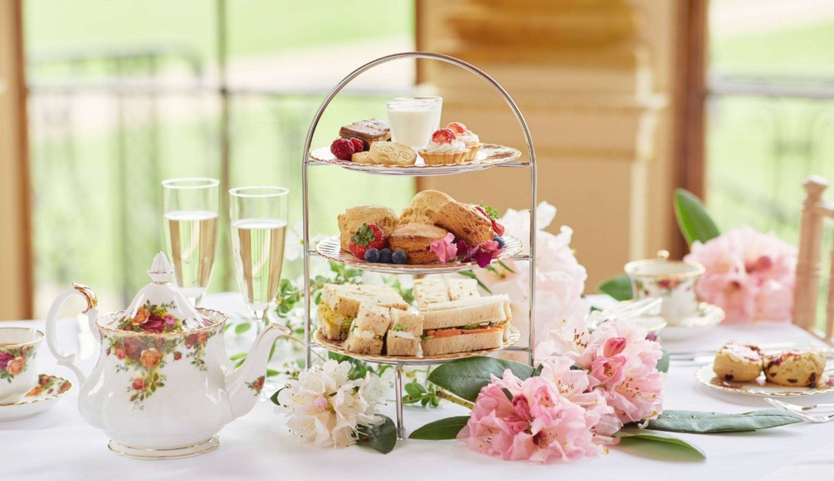A table is laid for afternoon tea with a white china patterned teapot and a cake stand with sandwiches and cakes in the centre. There are two glasses of champagne and a buttered scone with flowers on the table.