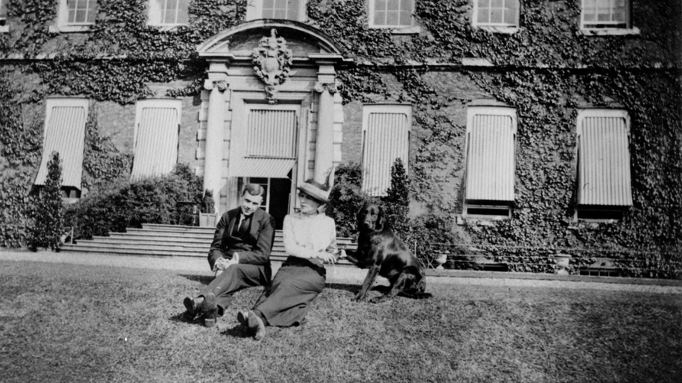 Historic black and white image of a man and woman and dog sat on the lawn in front of a grand doorway