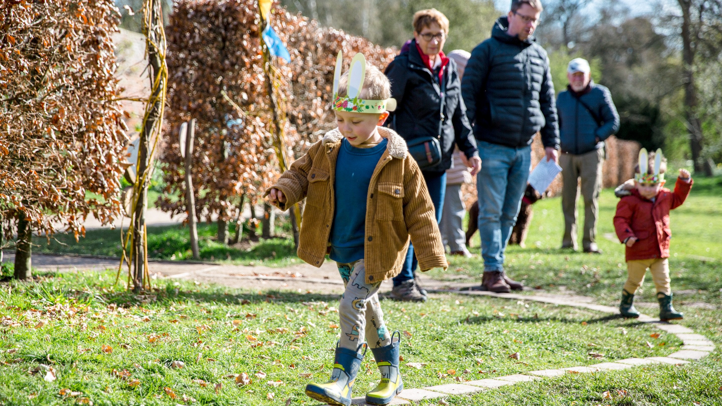 Young boy in paper bunny ears walking ina garden ahead of his family