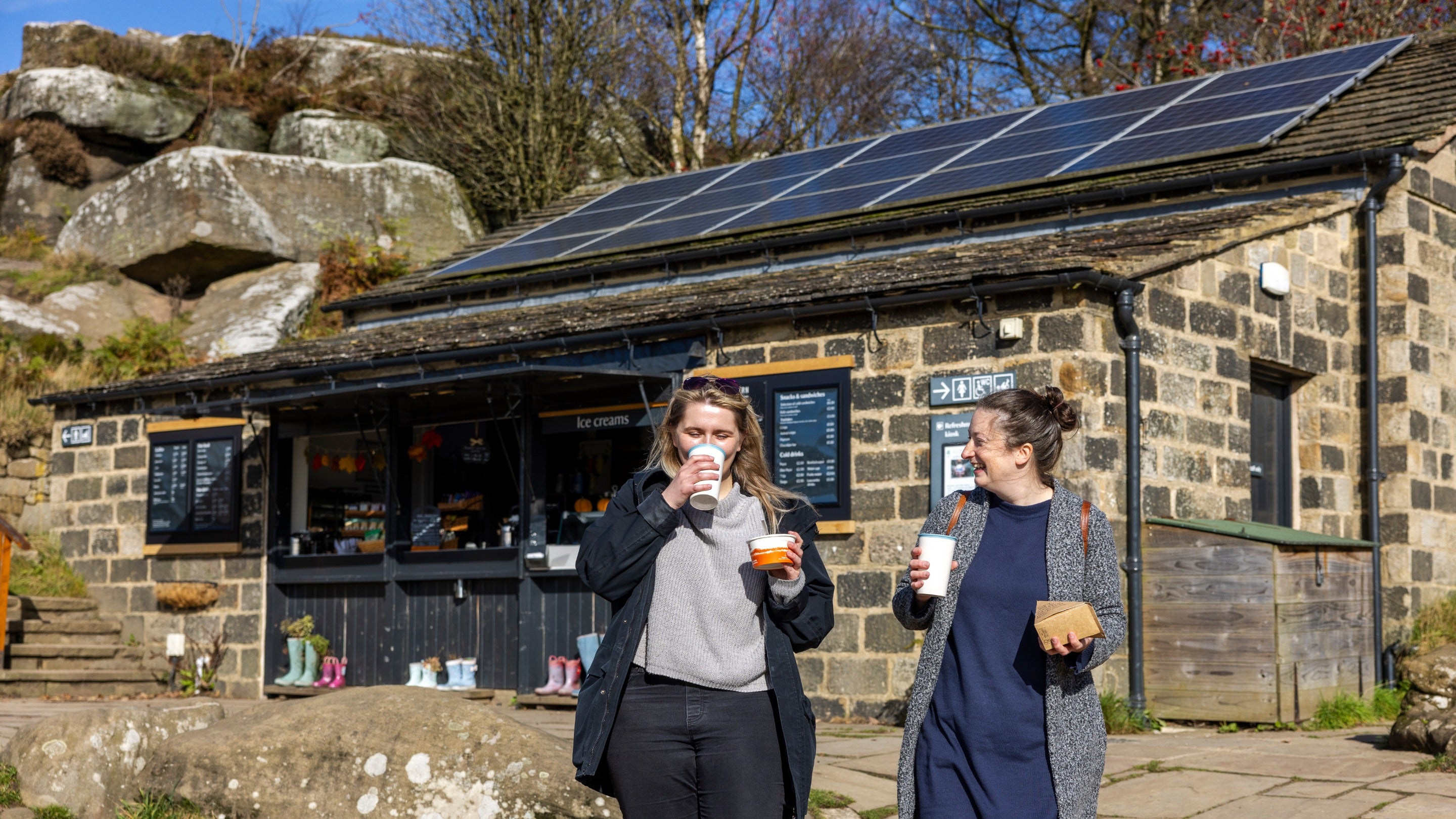 Two women walk away from a stone-built cafe enjoying a warm drink as they go with blue autumnal skies above