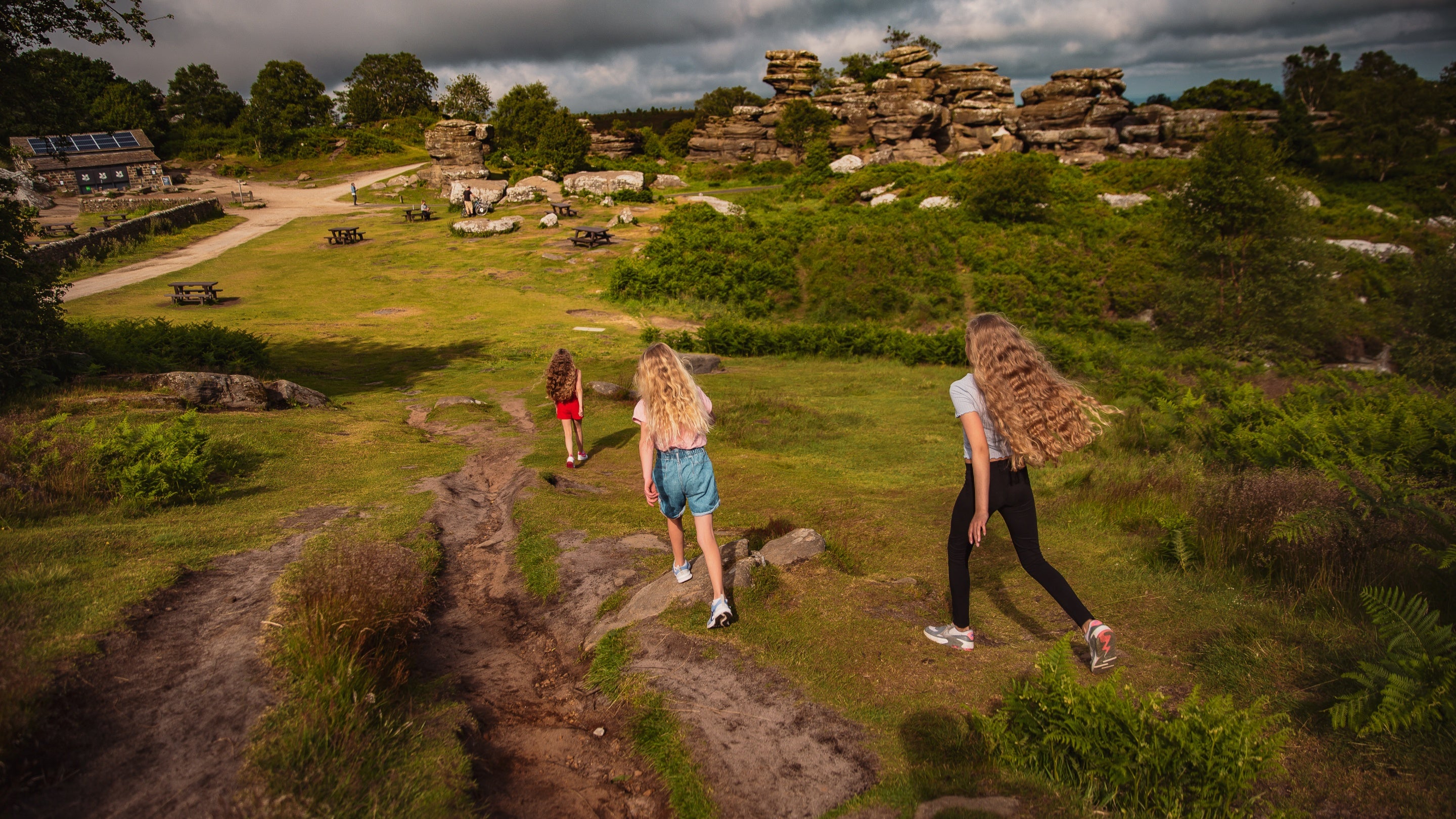 Three girls walk at Brimham Rocks with with their backs to camera down a part towards the rock formations in front of them