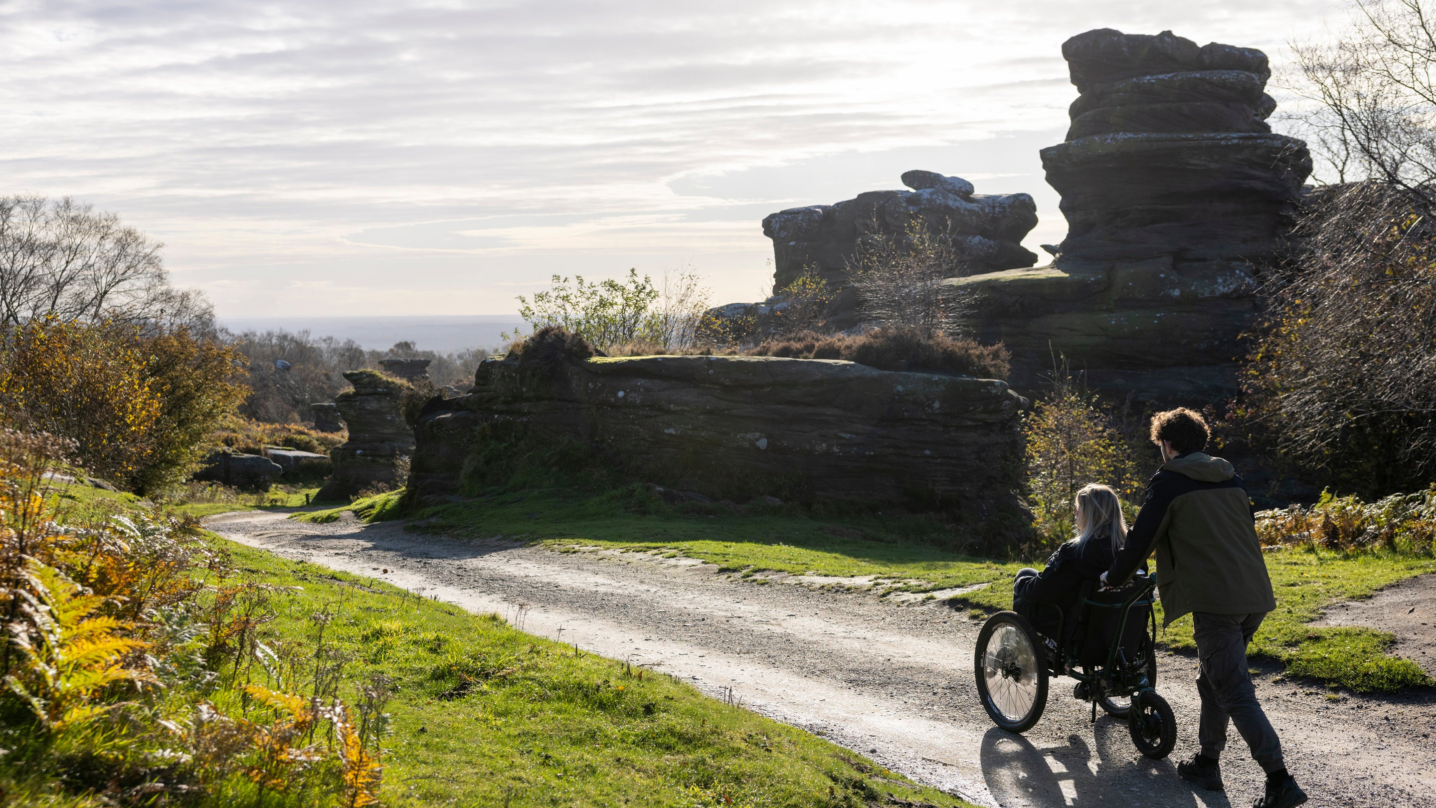 Visitor being pushed in a wheelchair along a wide gravel path at Brimham Rocks, with distinctive rock formations, low vegetation and open countryside stretching into the distance.
