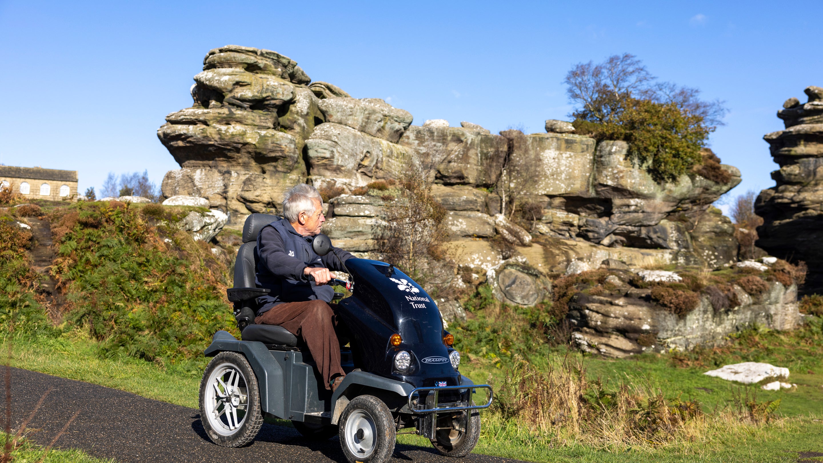 Visitor using a mobility scooter on an accessible tarmac path at Brimham Rocks, with large weathered rock formations and autumn vegetation in the background under a clear blue sky.