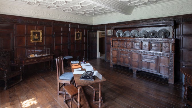 A dark panelled room with a white plaster ceiling and dark, period furniture.