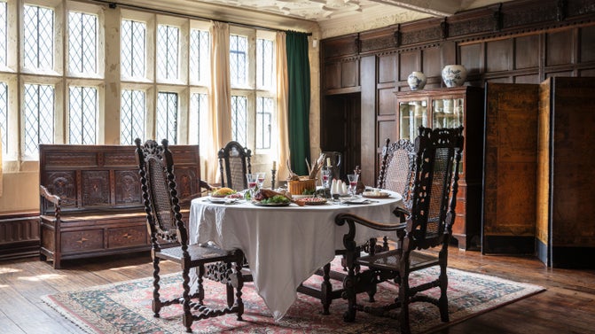 A dark wooden table stands in the centre of a room with dark panelling and mullion windows in the background