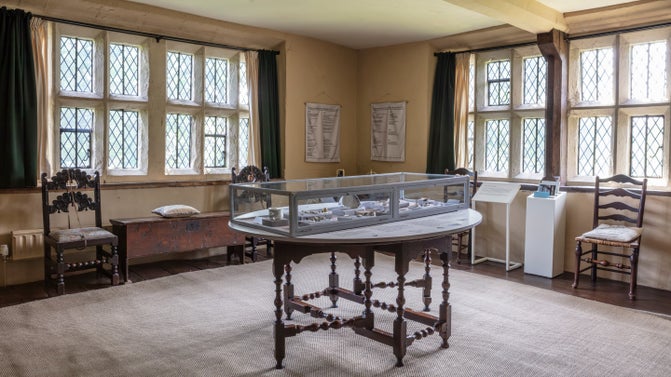 A table with a glass case on top stands on the centre of a room with mullion windows in the background