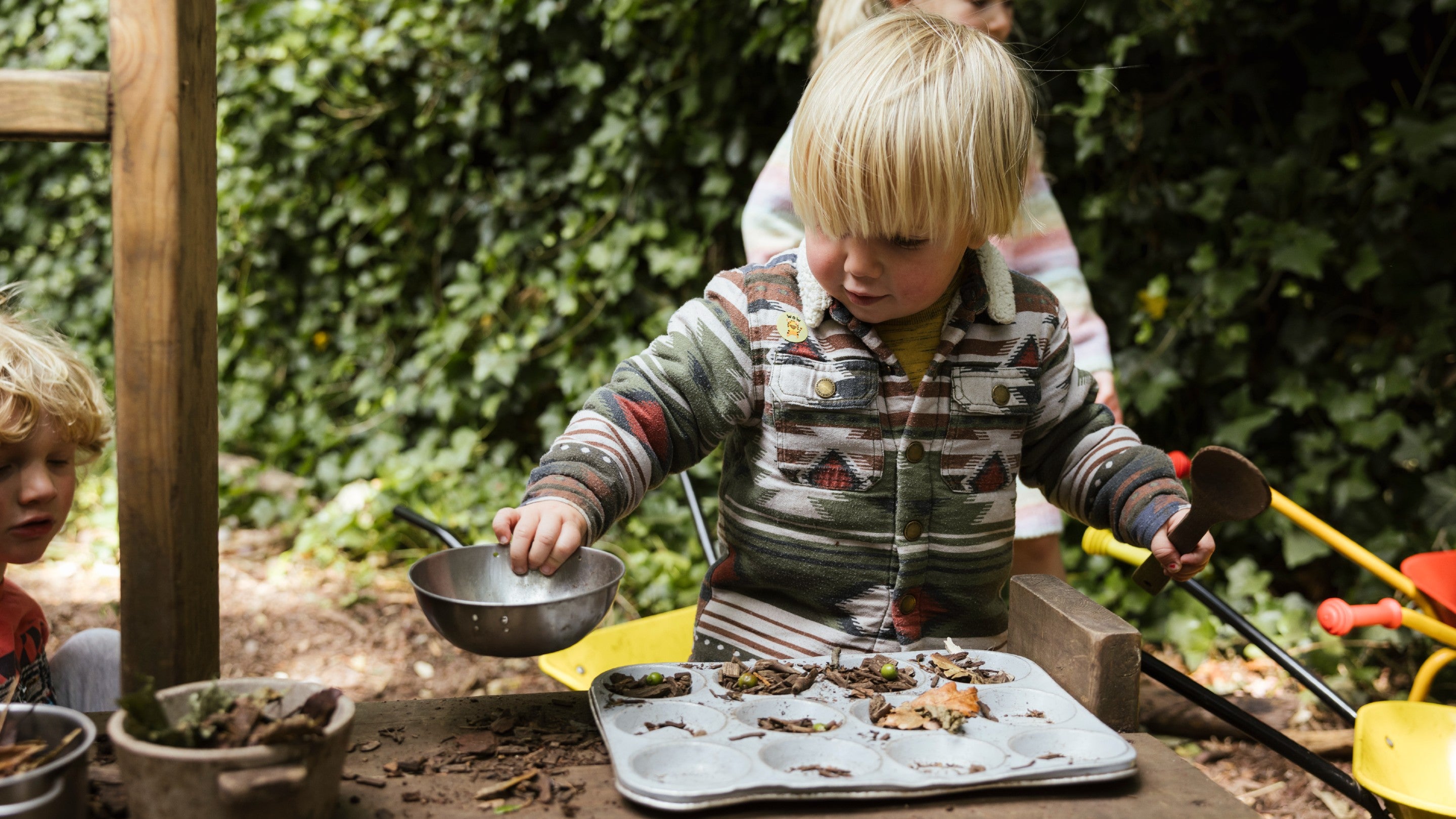 A boy in a knitted jumper is busy playing in the mud pie kitchen, concentrating on what he's doing.