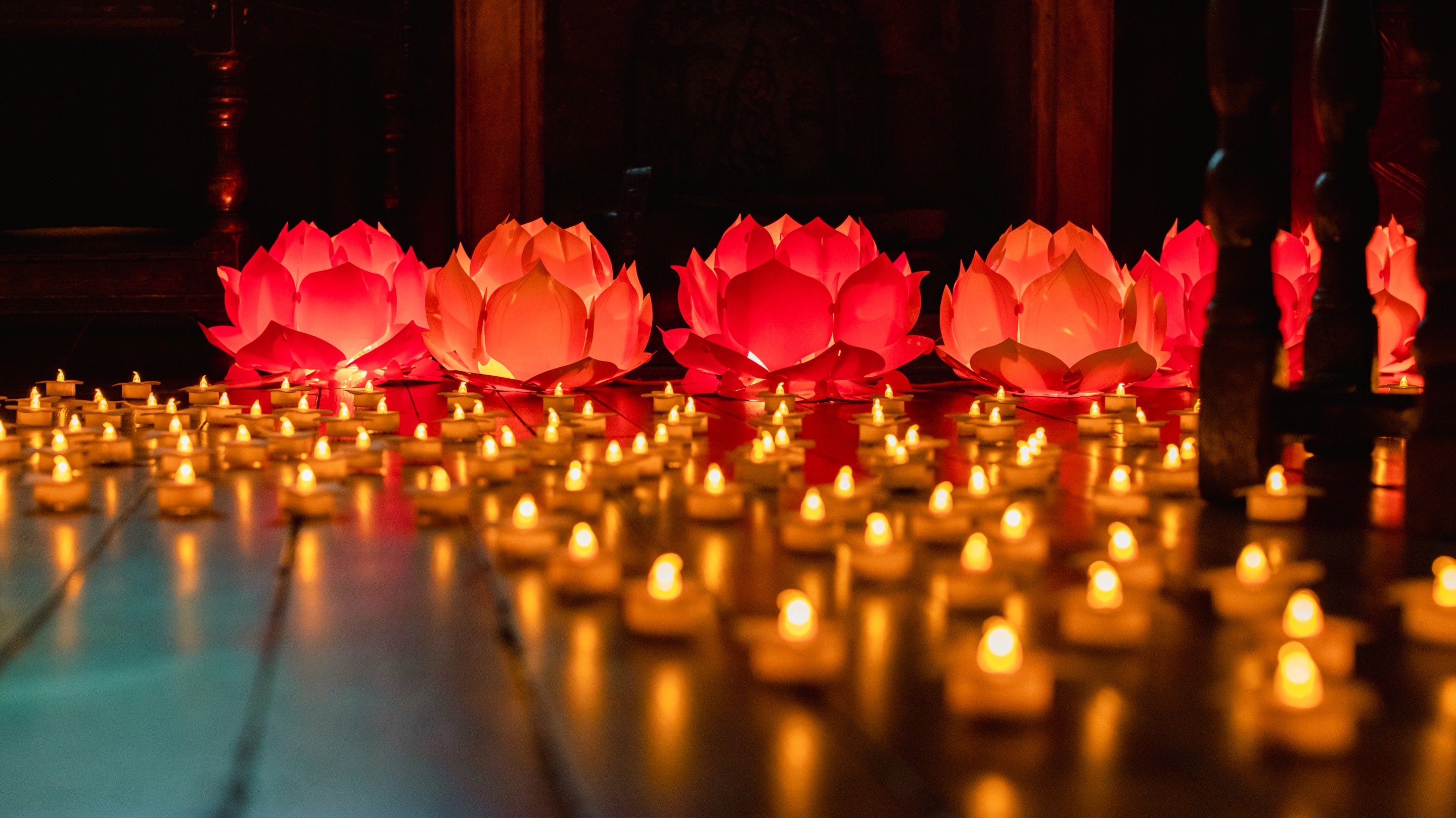 Orange glowing candles are arranged in circles on a dark wood floor with pink lit lotus flower displays in the background