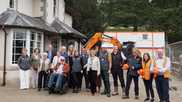 National Trust staff, project partners and contractors gather outside Studley Royal's visitor facilities to celebrate the start of construction on the Studley Revealed project, with an excavator visible behind the group.
