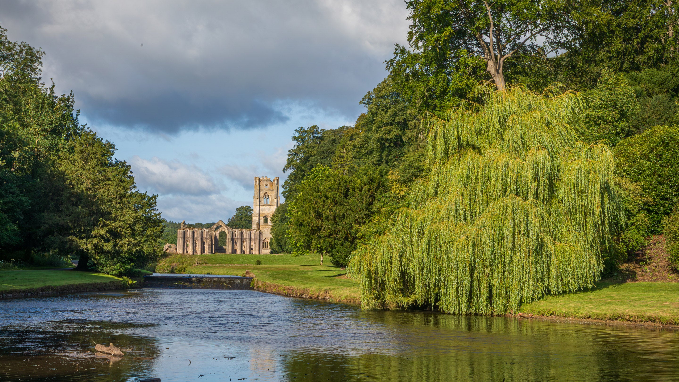 Ruins of an abbey seen in the distance beyond a bend in a river, with a large willow tree and grassy banks in the foreground under a cloudy sky.
