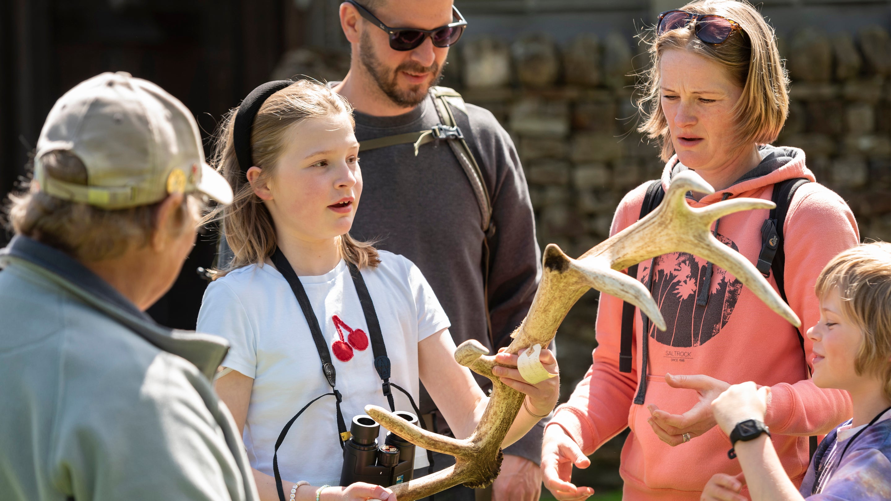 A small group of people stand outdoors examining a large deer antler. One child holds the antler while the others gather around to look closely.