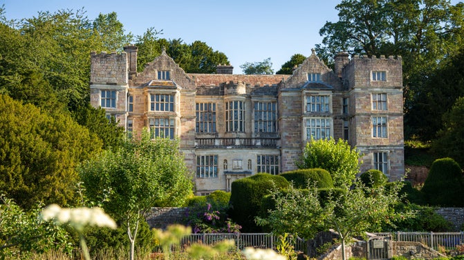 Fountains Hall, a historic stone house, seen beyond a garden with trees and neatly trimmed hedges on a sunny day.