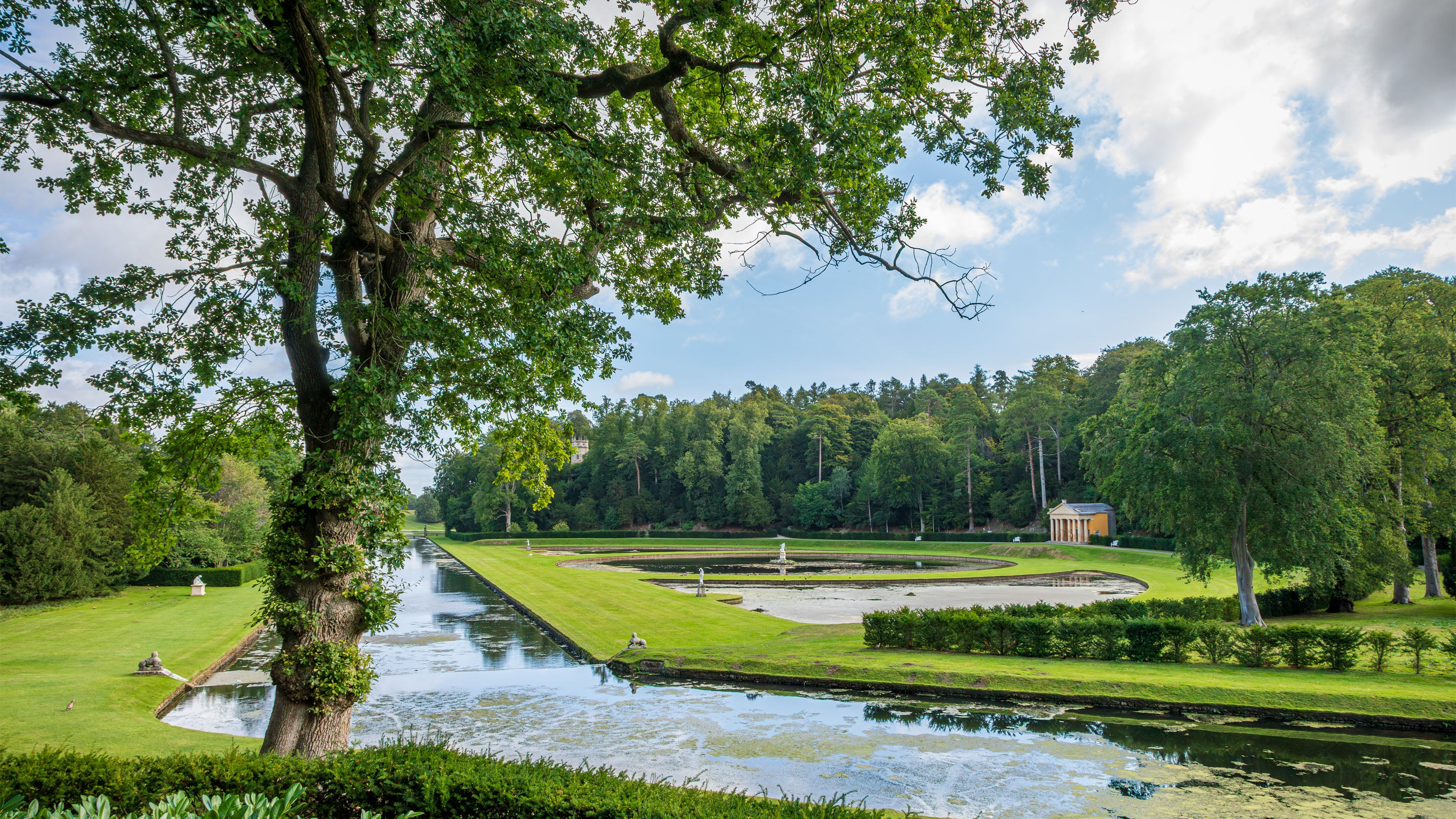 Studley Royal Water Garden with a straight canal, circular pond and lawns, framed by trees, with a small classical temple in the distance under a partly cloudy sky.