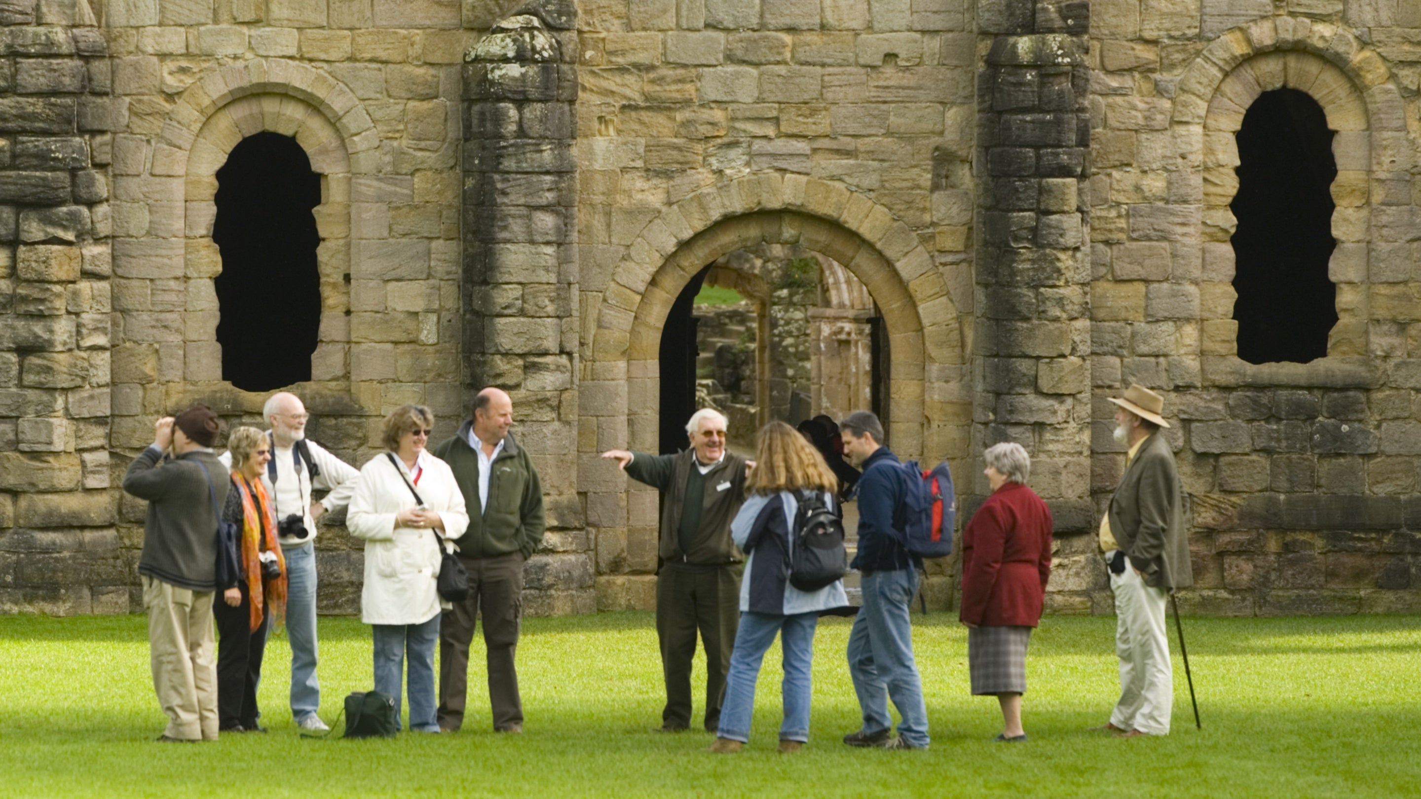 A group of visitors are listening to a guide as they stand on the grass in front of the ruins of Fountains Abbey, North Yorkshire.