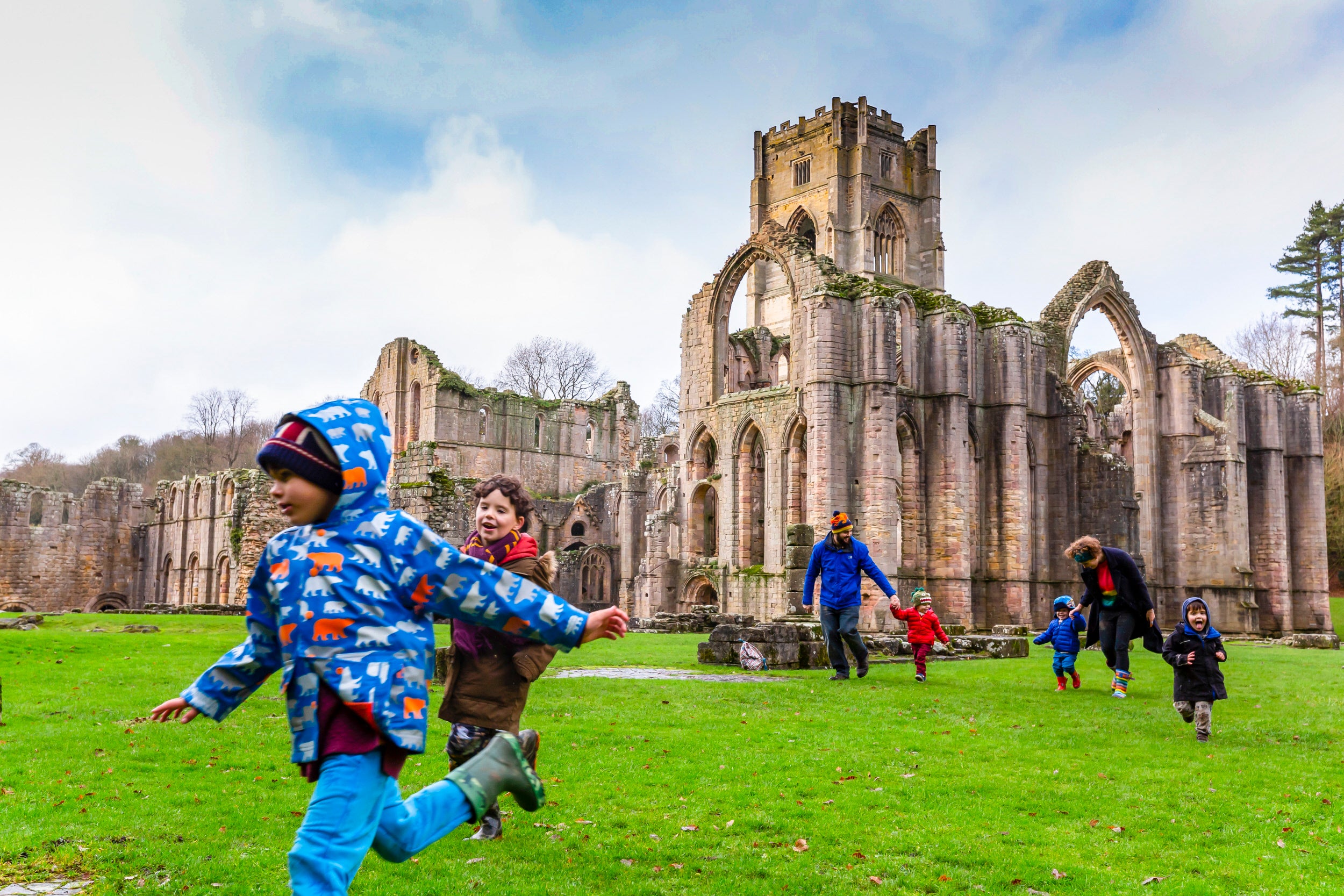 A family running across an open green space, with dramatic stone ruins behind