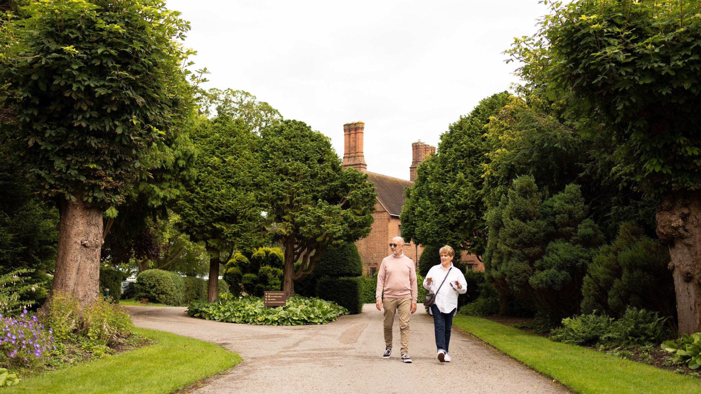 Two people walking down a long drive with trees either side and chimneys seen in the distance