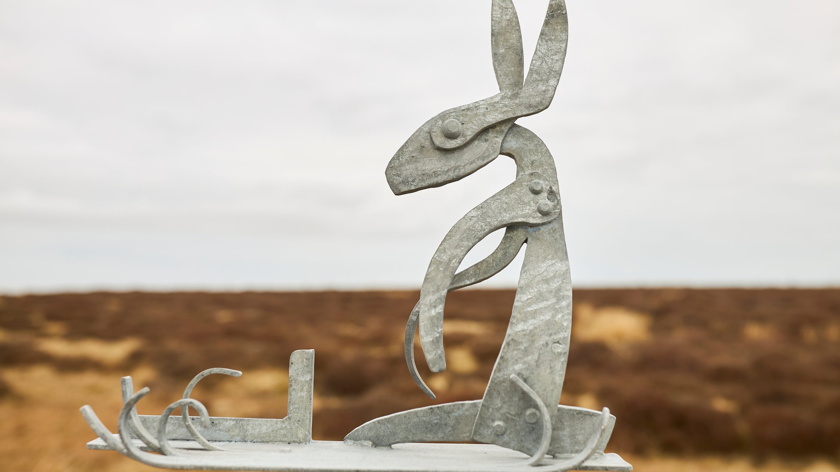 A metal sculpture of a hare sitting upright with elongated ears and abstract, curved limbs. The sculpture is set against a blurred background of an open landscape with brownish vegetation and an overcast sky.