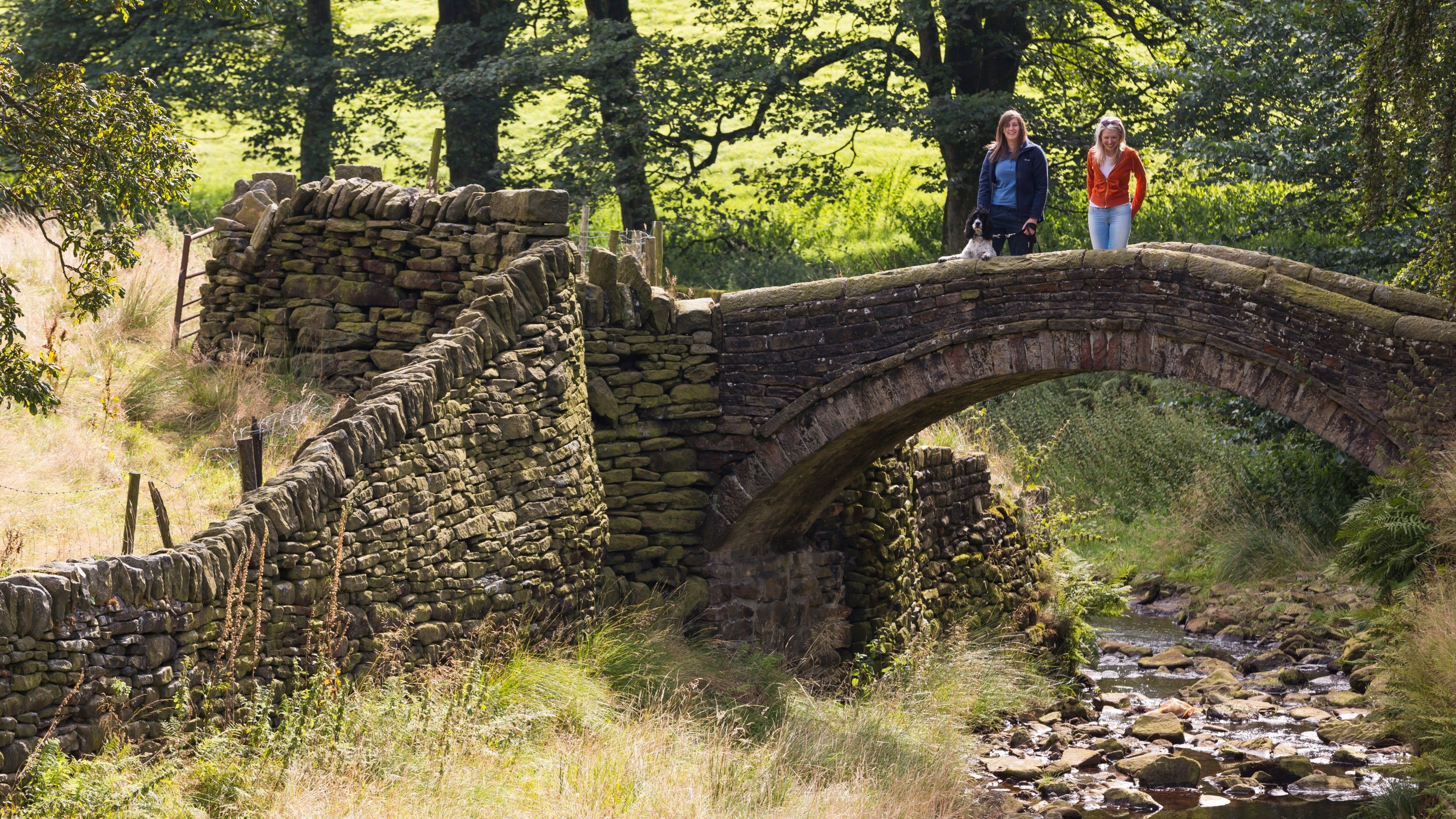 Two women stand on Eastergate bridge, on a sunny day. The water levels are low in the beck below them