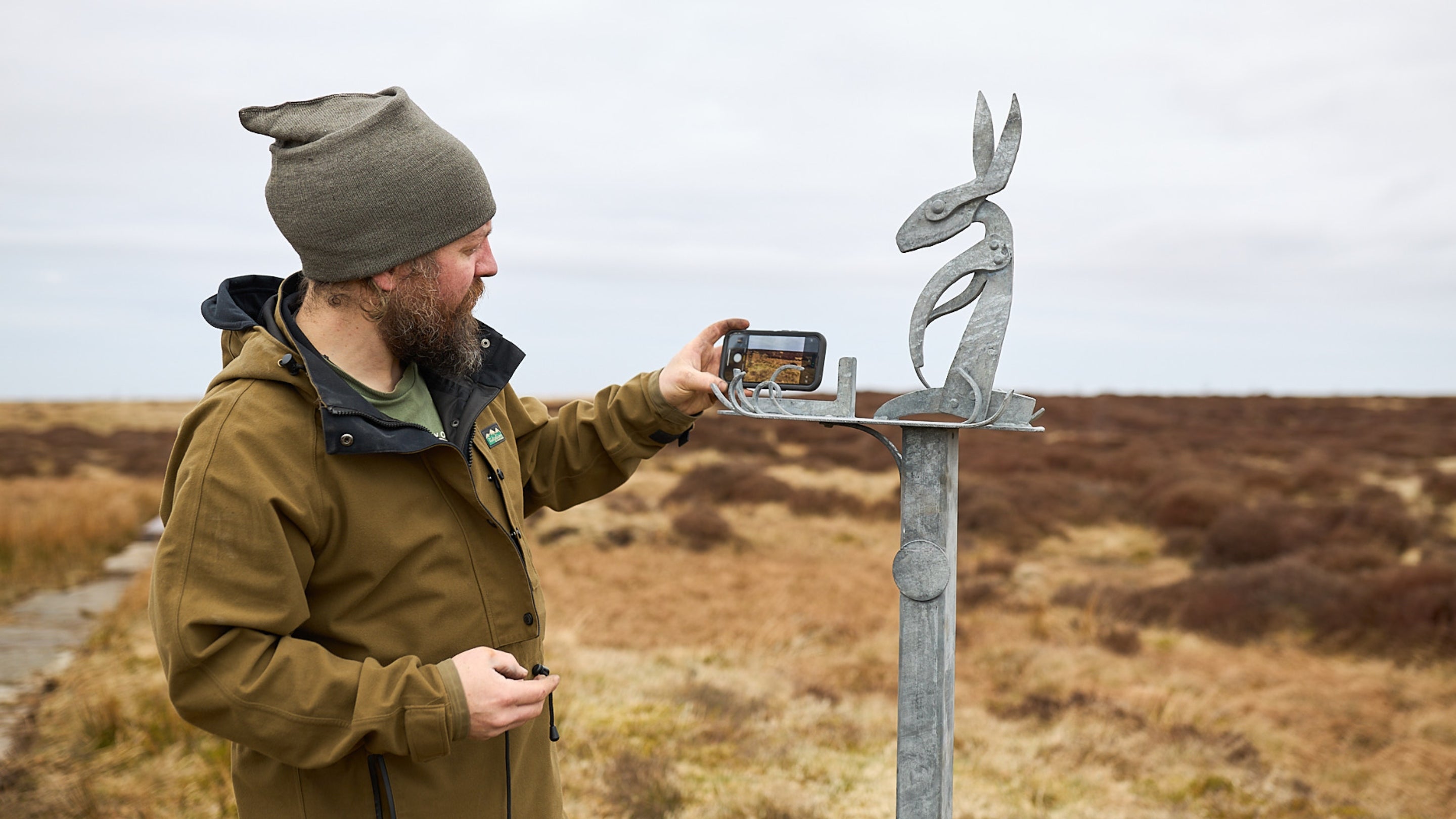 A person in a brown jacket and gray beanie is taking a photo with a smartphone of a metal sculpture of a hare. The sculpture is mounted on top of a wooden post in an open, grassy landscape with brown vegetation and cloudy skies