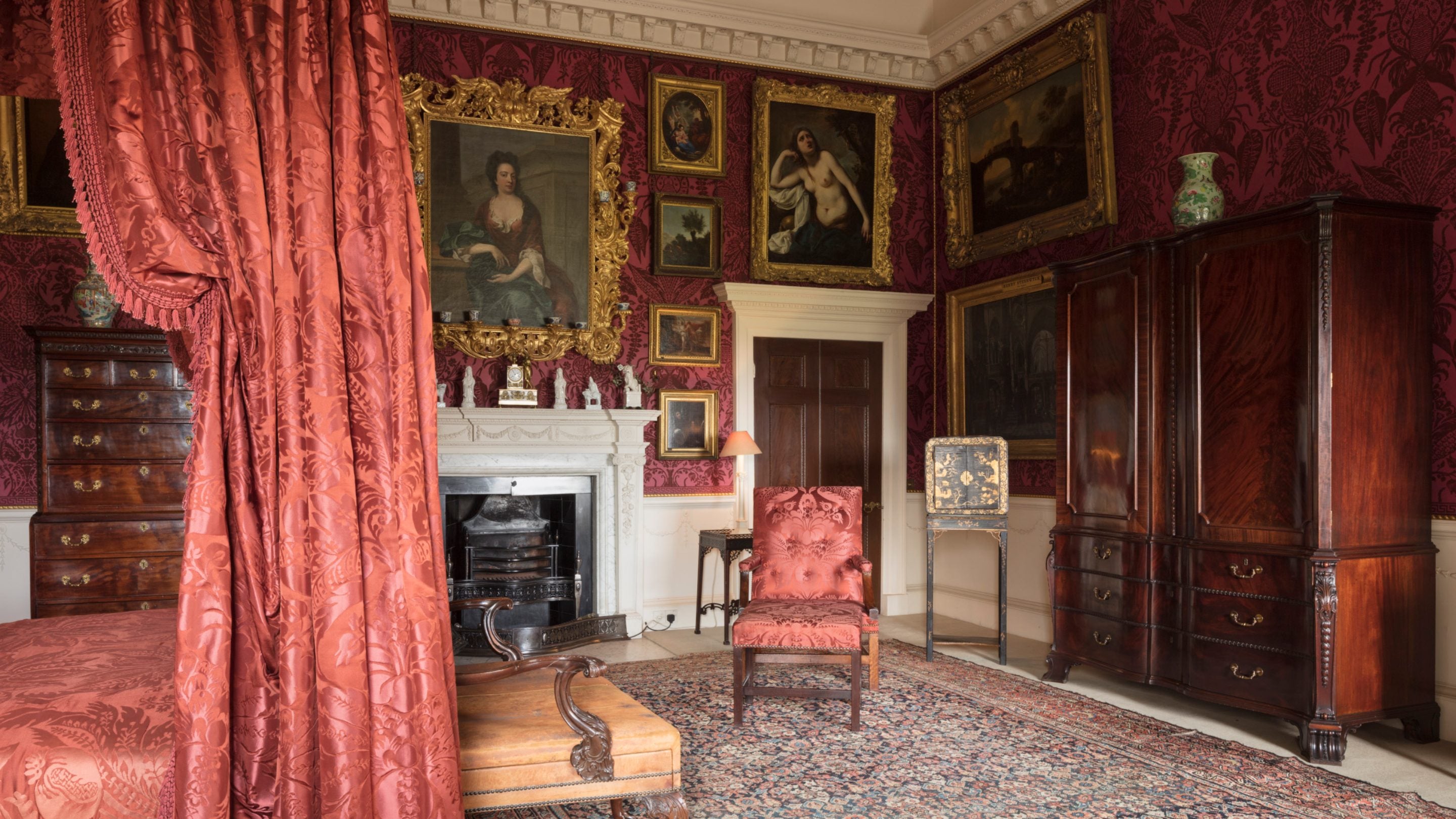 An ornate, richly decorated bedroom, with a four-poster hung with red damask, mahogany furniture and classical portraits on the walls