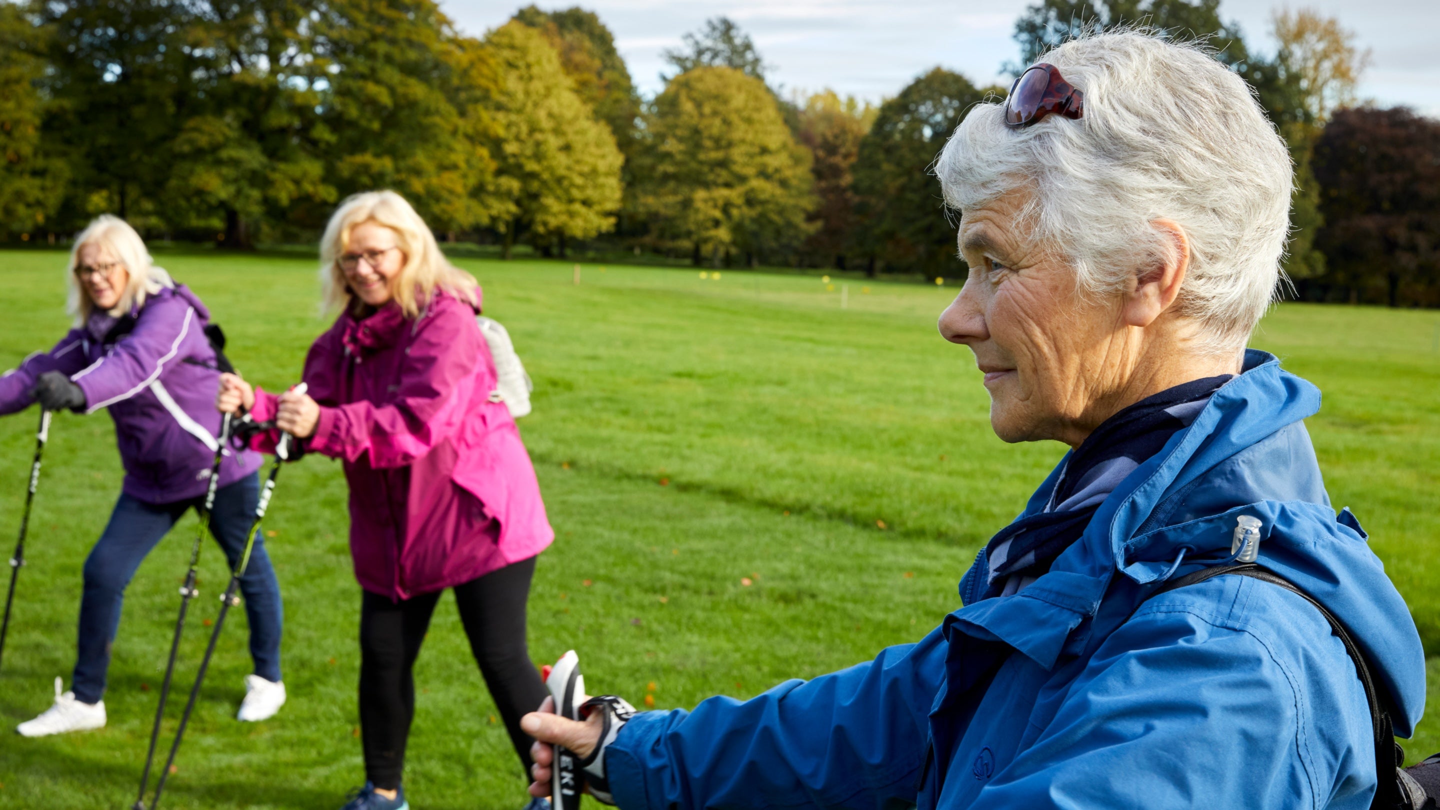 Three women in a grassy field step forward to lean on their walking poles, which they have in each hand.