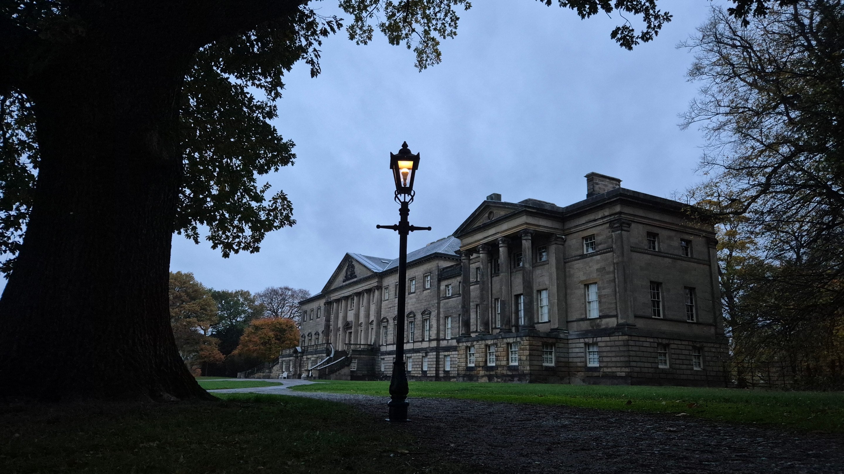 Looking through the trees at dusk at Nostell house in Winter. There is an old fashioned lamp post in the foreground which is lit.
