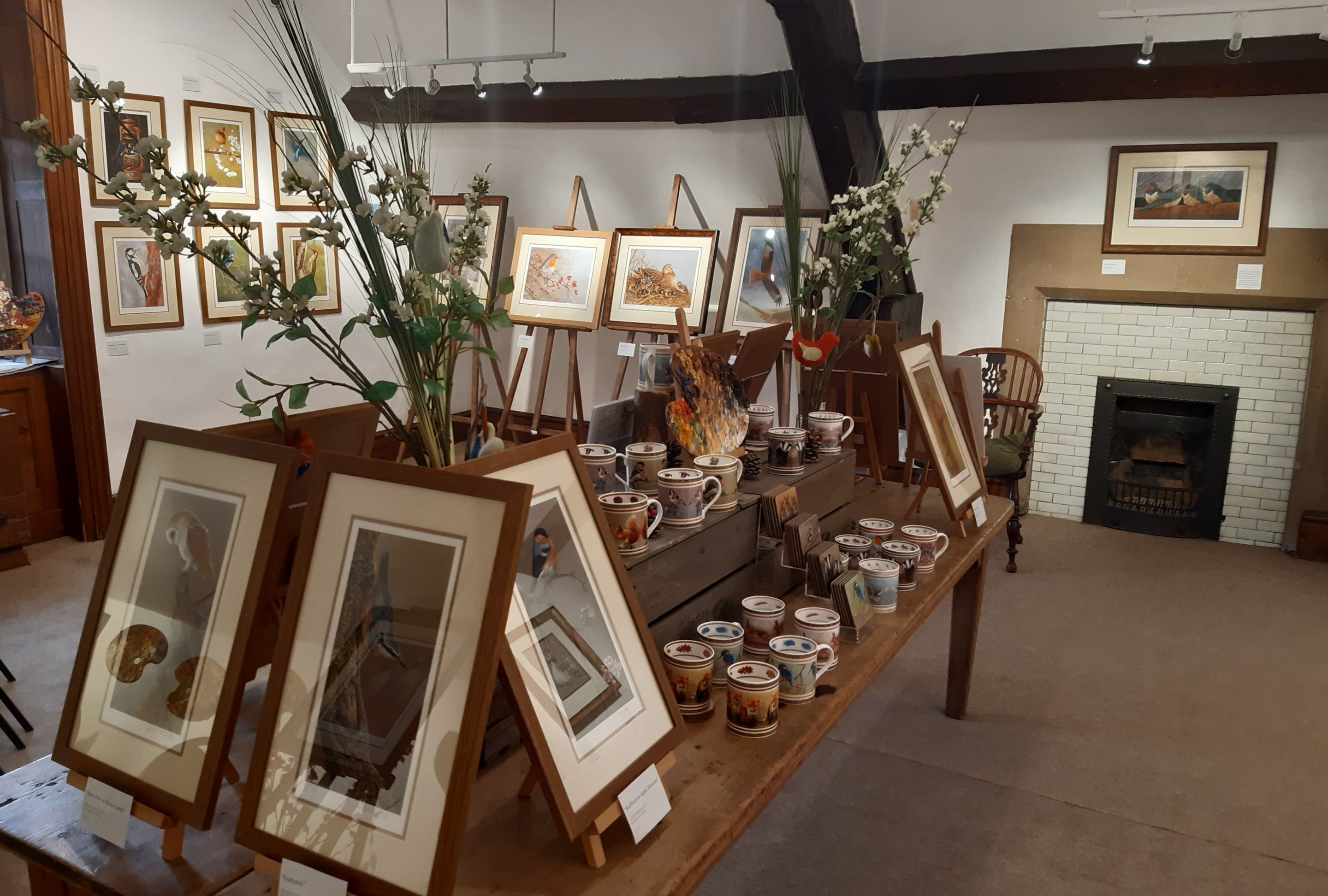 Framed artwork on the walls in a wooden-beamed room. There are  framed works on easels and mug and flower displays on a table in the centre of the room.