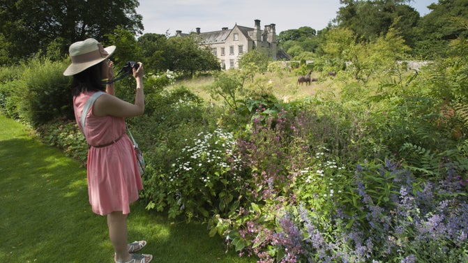 Woman in summery clothes taking photos of a flower border with a manor house in the distance