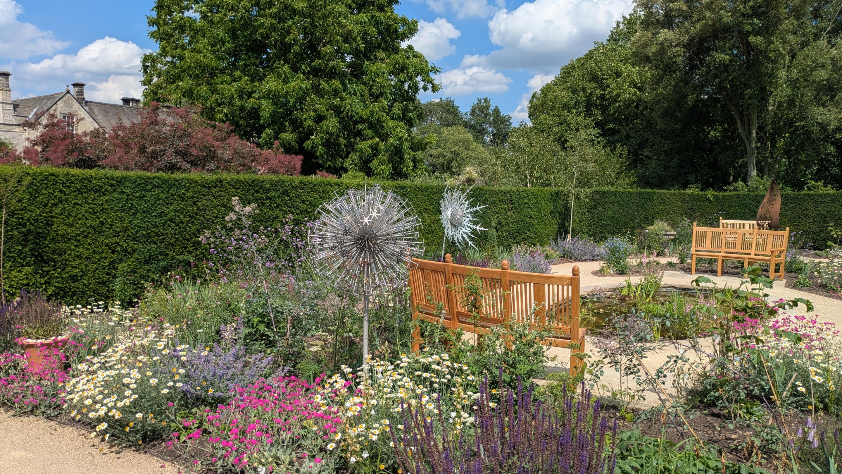 View across a colourful garden with sculpture, paths, benches and a pond