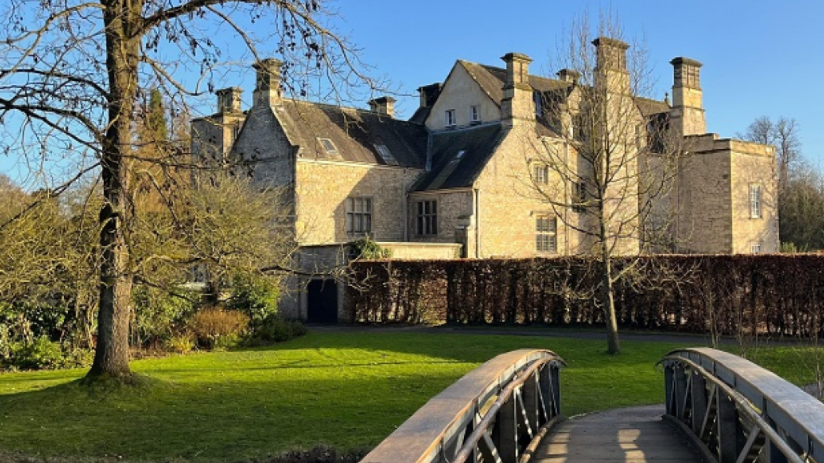 View across a bridge to a winter hedge and trees and large manor house behind