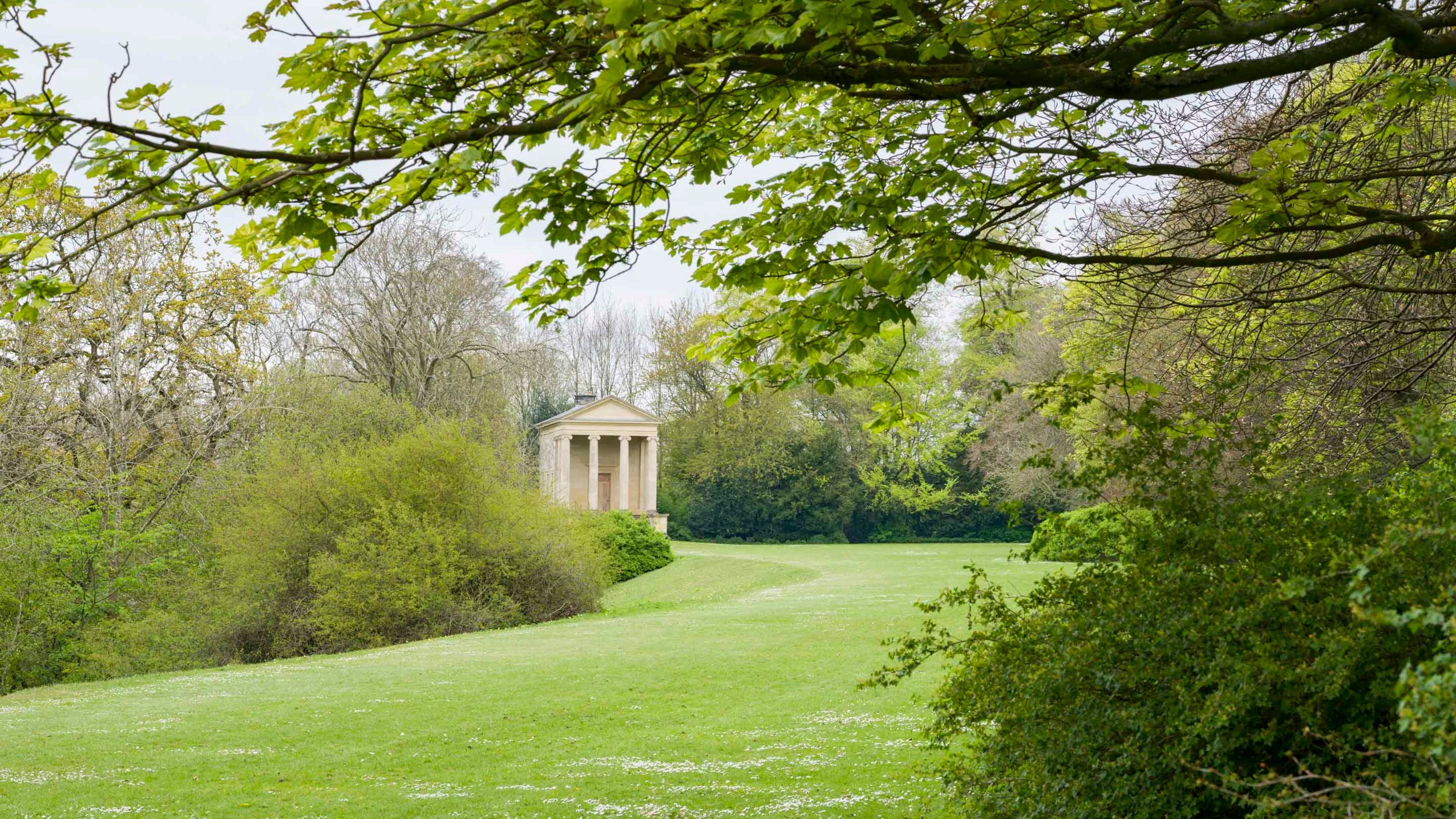 A view down the lawn of the Terrace at Rievaulx Terrace, North Yorkshire, with the Ionic Temple in the middle ground; in the foreground are mature trees and bushes