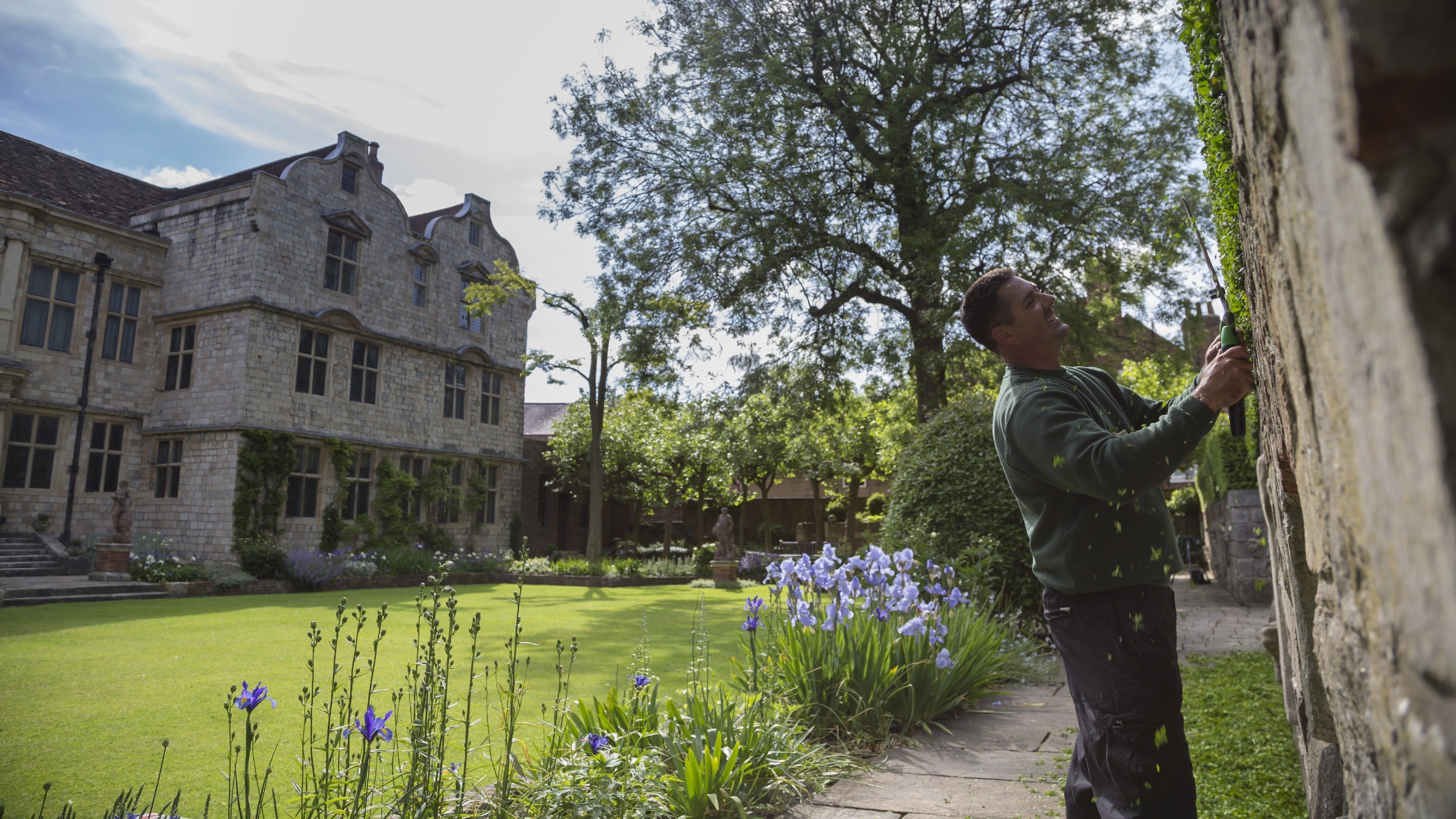 Man using handheld hedge cutters with a lawn and stone house visible behind