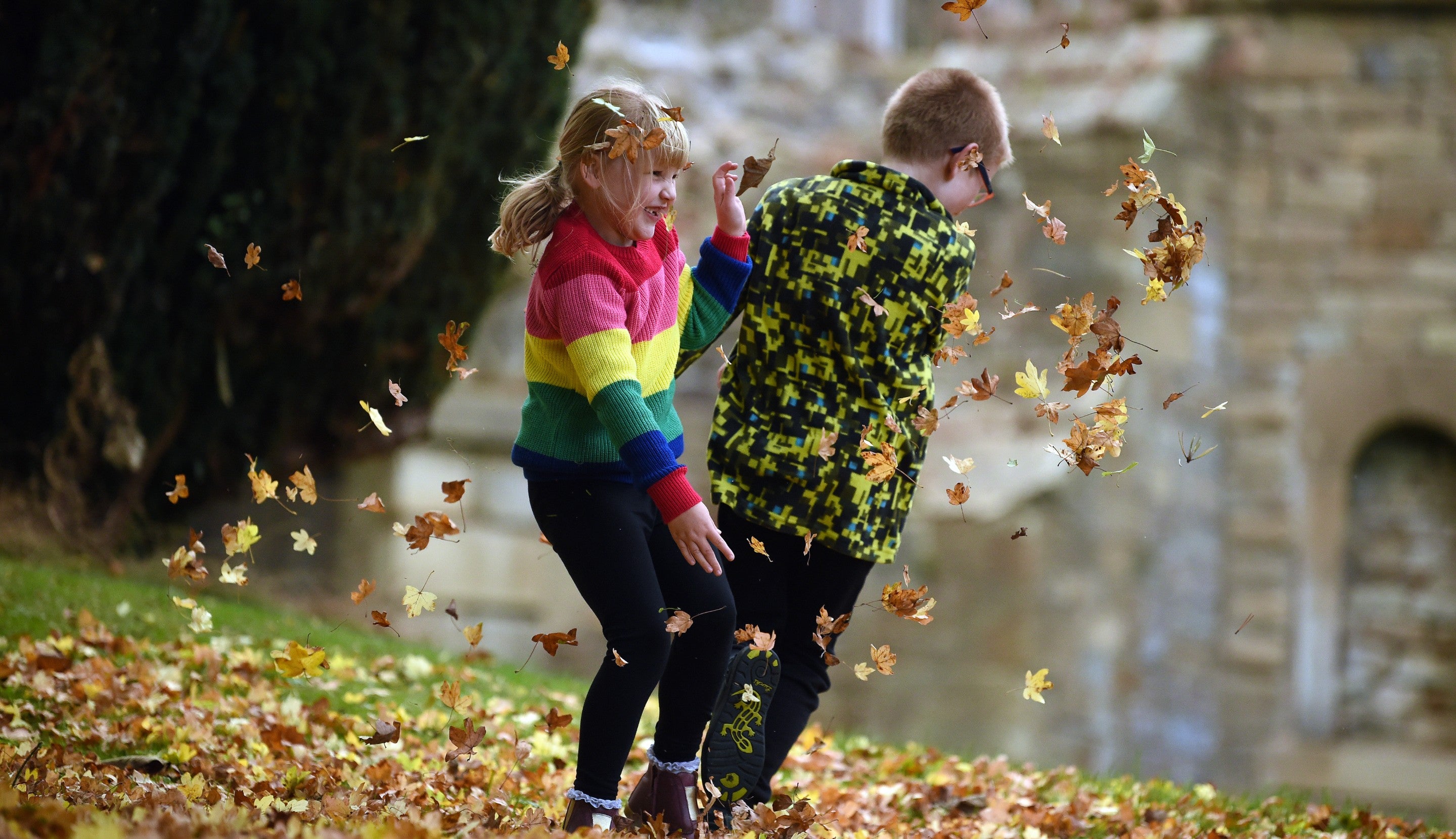 A girl in a rainbow striped jumper and a boy in camouflage jacket kick and throw leaves at each other